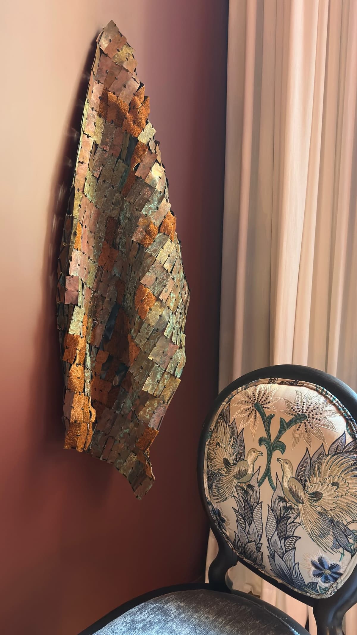 A hand-forged copper wall sculpture against an oxblood wall, beside an embroidered occasional chair.